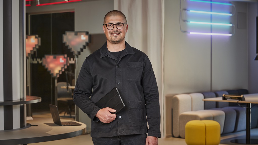 Man klädd i svarta kläder håller sin dator under armen. Står på ett kontor med häftig bakgrund 